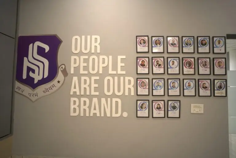 Here is a **125–140 character SEO-optimized alt text** for this image: ### ✅ **ALT Text (125–140 characters)** *Wall display at an MBA college featuring the slogan “Our People Are Our Brand” with framed faculty and achiever photos highlighting campus excellence.*